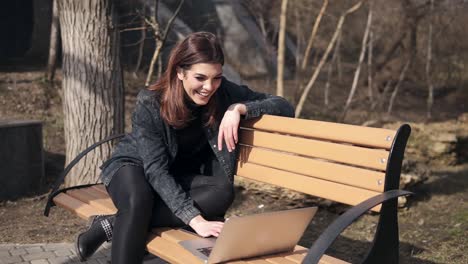 Young-attractive-brunette-girl-is-sitting-on-the-bench-in-park-waving-at-someone-in-her-laptop,-chatting,-laughing-and-having-fun-time.-Outdoors-footage