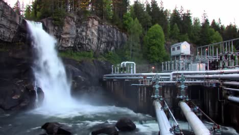 a beautiful waterfall flowing next to an industrial water processing plant surrounded by lush greenery and rocky terrain at dusk