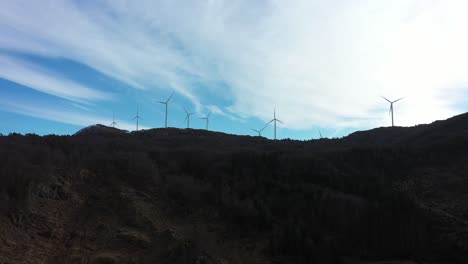 Wind-turbines-at-mountain-Midtfjellet-in-Fitjar-Island-in-Norway---Sideways-moving-aerial-with-dark-foreground-and-silhouette-wind-turbines-against-bright-blue-sky-with-fast-moving-clouds