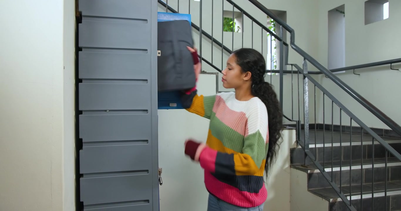 Girl descending stairs, approaching blue lockers, opening locker, retrieving black bag for class