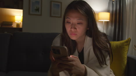 anxious woman spending evening at home sitting on sofa reacting to internet or social media news message or story on mobile phone