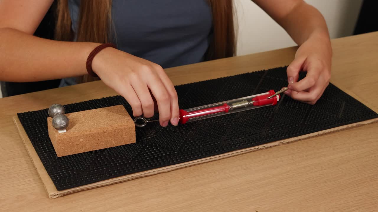 Hands use a spring scale to measure frictional force on a block. Indoor setting, close-up view, natural lighting