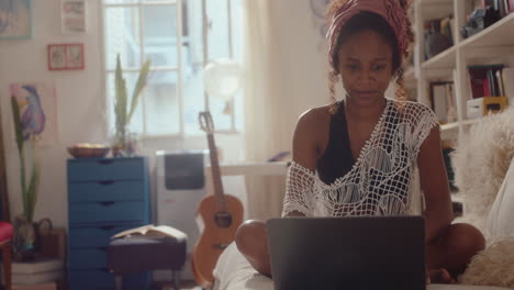 black girl sitting on sofa at home and online messaging on laptop