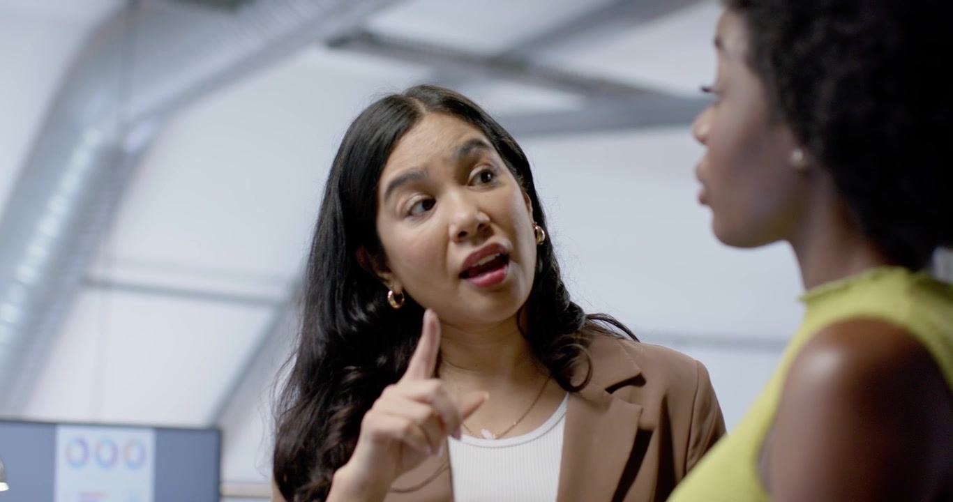 Female staff in office reviewing laptop, brown blazer clarifying point while yellow top listening