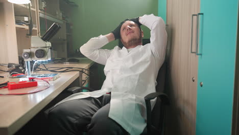 a middle view of a relaxed technician in a white lab coat leaning back in his chair, hands behind his head, taking a break from working with electronic components in a lab setting