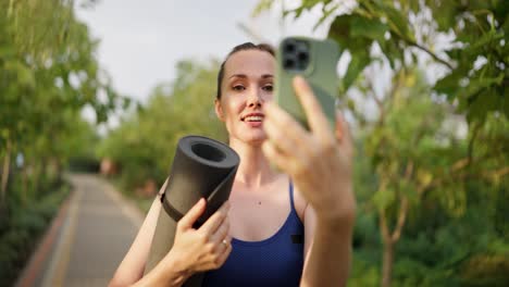 woman taking a selfie video call while holding yoga mat in a park