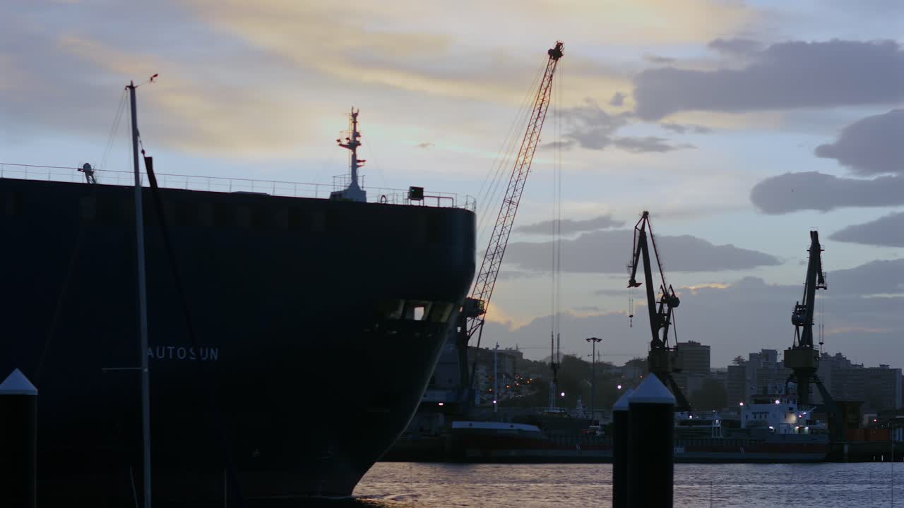 Large vehicle carrier ship departs Pasaia Bay, Spain, with port cranes visible in the background. Captured from a side angle