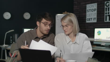 Young-business-man-and-woman-working-together-front-laptop-in-dark-office