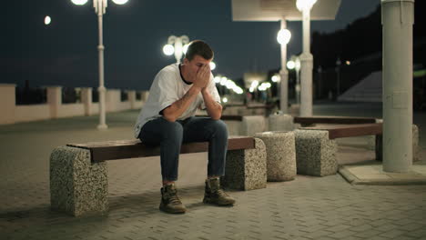 tired man sitting on bench at dusk, deep in thought, with street lights and urban structures in background, he appears stressed or reflective in city setting at night