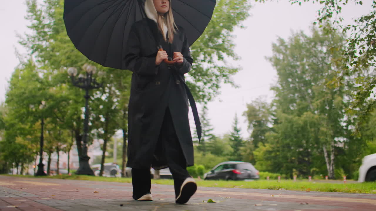 Young woman in long black coat and hood holding umbrella walking on tree-lined pathway with fallen leaves, cars and buildings in background, calm rainy day urban lifestyle moment