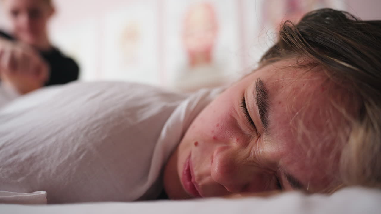 Client lying down with cheek pressed against soft surface while receiving gentle face massage, eyes open in calm expression, therapist s hands slightly blurred in background, wellness posters on wall