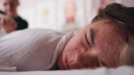 client lying down with cheek pressed against soft surface while receiving gentle face massage, eyes open in calm expression, therapist s hands slightly blurred in background, wellness posters on wall