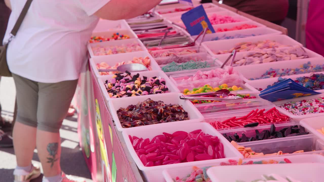 Adult examines assorted gummies and sweets at vibrant outdoor confectionery stall, bright daylight, static shot
