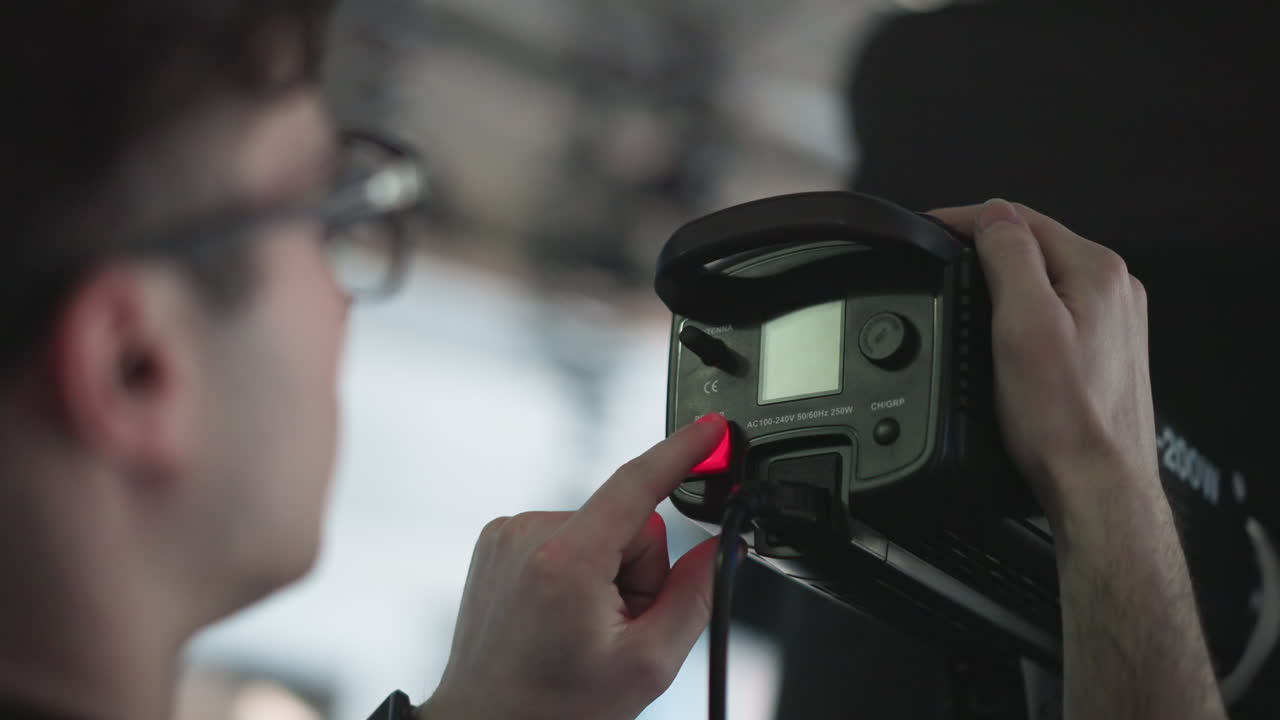 Camera technician pressing power switch on LED studio light control panel, adjusting settings during broadcast shoot with cables and mounting hardware visible in professional production environment