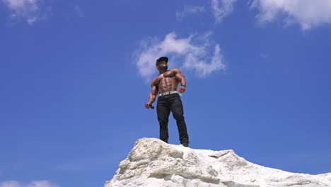 Bare torso of black skinned athlete. African american bodybuilder on the top of white hill against blue sky background. View from below.