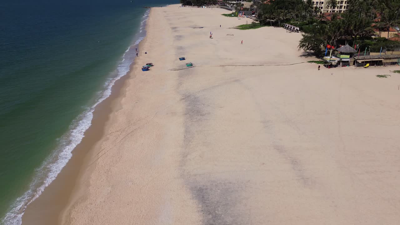 Drone flies forward over Mui Ne beach, panning up from bird’s-eye view to reveal jetskis parked on white sand, seaside on the left, and resorts lining the shore on the right.