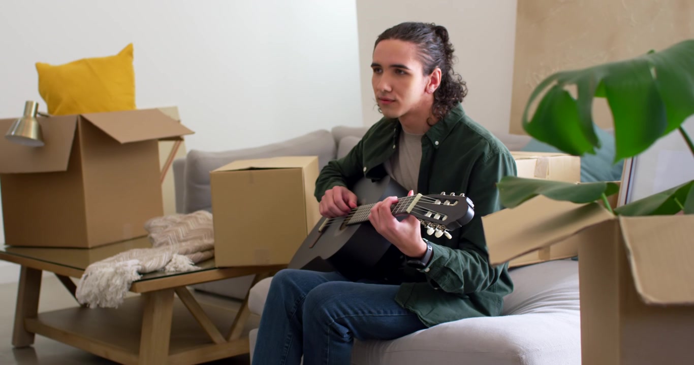 Homem afro-americano no sofá entre caixas de mudança, pegando um violão acústico e tocando, se acomodando.