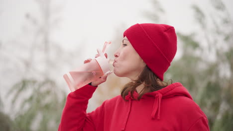 lady in red beanie and hoodie sipping water outdoors with pink bottle, set against blurred greenery background in foggy atmosphere