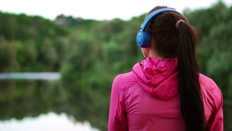 a girl in a pink jacket and blue headphones stands with her back and looks at the river early in the morning after a run