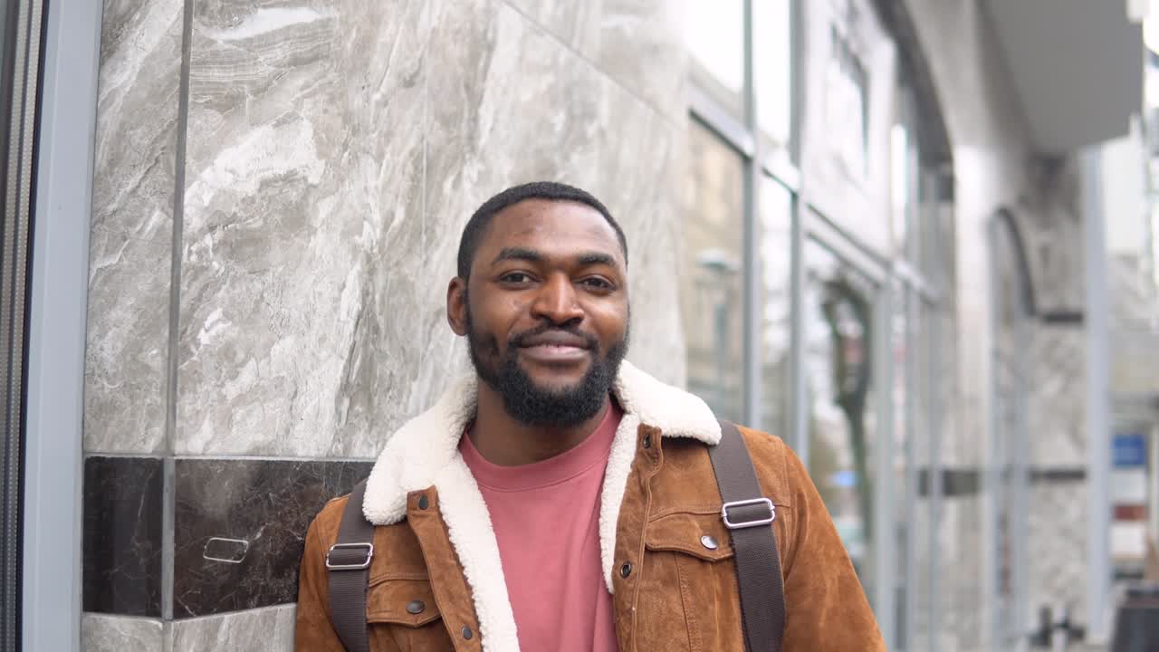 Bearded black with medical mask and shoulder backpack stands outdoor near the office center