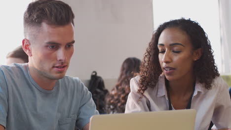 Female-Tutor-Giving-One-To-One-Support-To-Male-Student-Working-At-Desk-On-Laptop