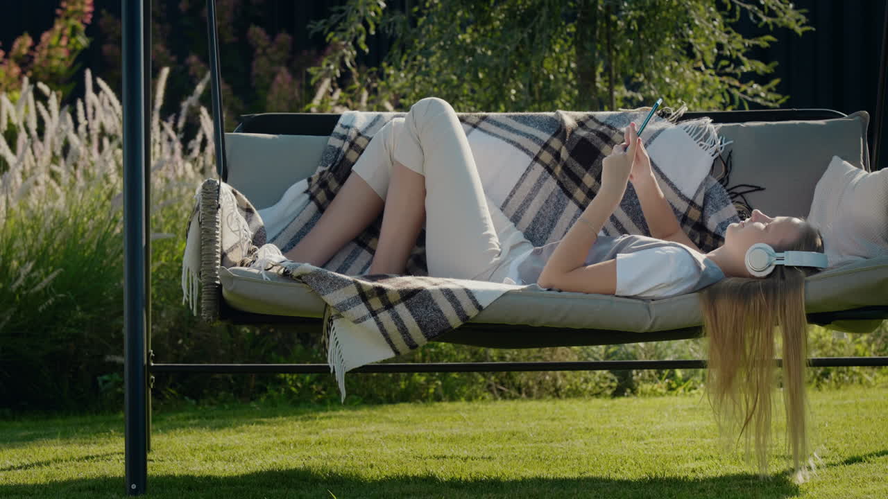긴 머리카락을 가진 십대 소녀는 헤드폰으로 음악을 듣고, 집 뒷마당의 정원 스에 누워 있습니다.