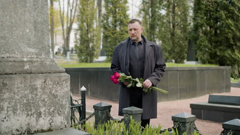 hombre con impermeable negro y traje sosteniendo rosas rojas frente a una lápida en un cementerio 3