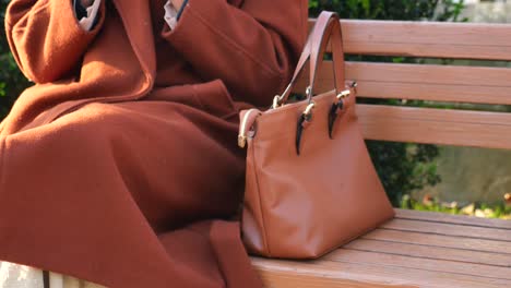 woman in a brown coat sitting on a bench with a bag