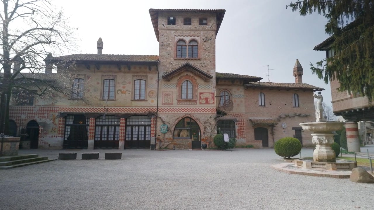 Un visitatore entra in un tranquillo cortile di Grazzano Visconti incorniciato da un arco, rivelando il palazzo neomedievale e la fontana centrale