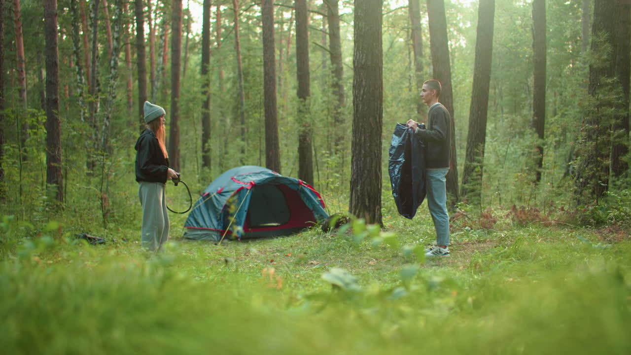 Young woman in knit hat watches man hold and spray tent fabric in sunny forest campsite with pitched tent behind them surrounded by tall trees and lush greenery