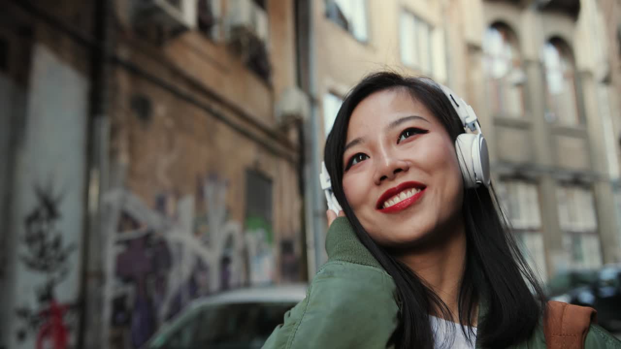 primer plano de la joven encantadora y feliz mujer con estilo en los auriculares que van a la calle en los barrios marginales y escuchan la música alegremente