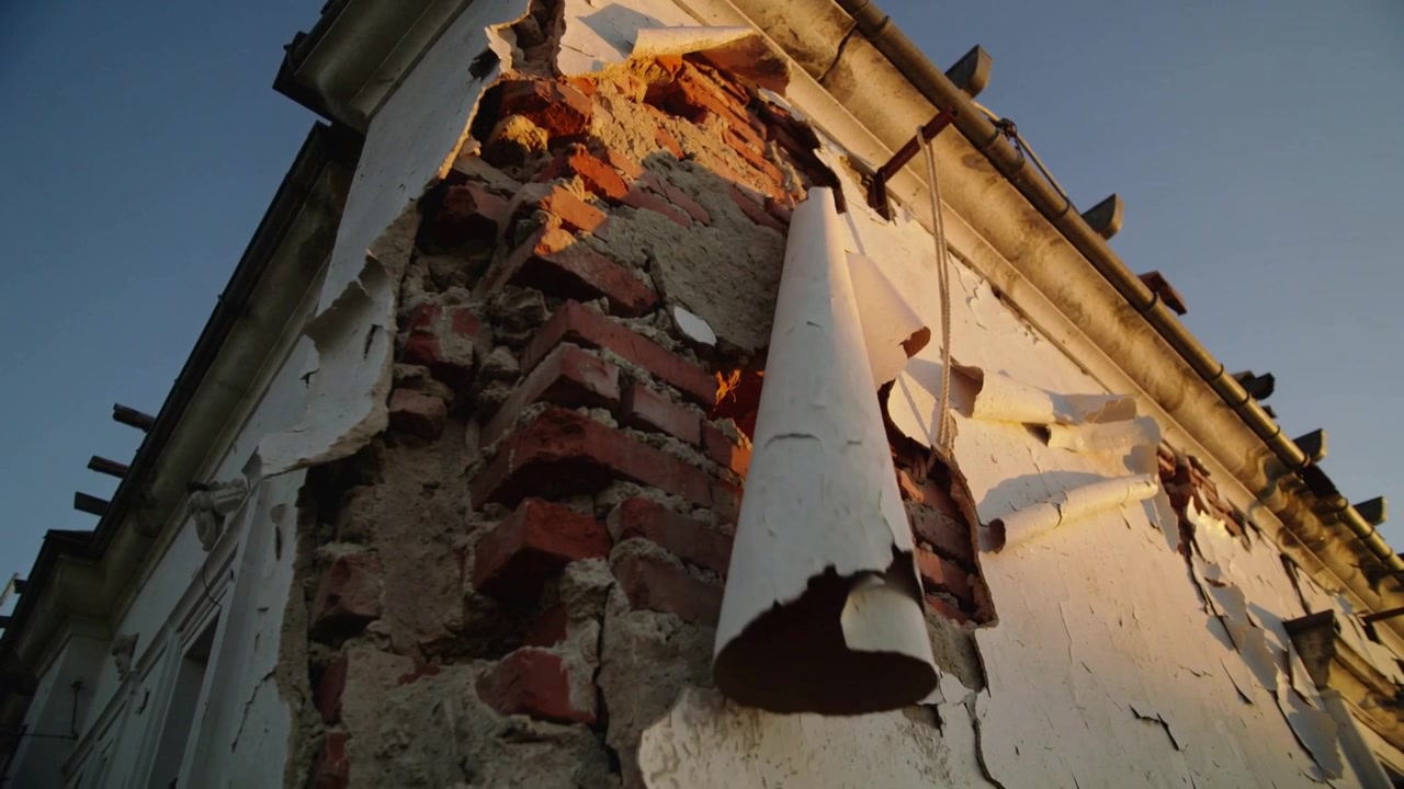 Telecamera inclinata che cattura l'angolo danneggiato alla linea del tetto, rivelando intonaco scrostato, mattoni rossi e grondaia.