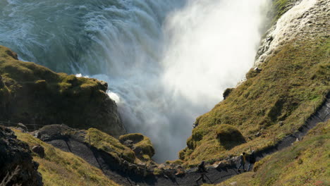 Close-up-shot-of-person-hiking-on-trail-above-gigantic-Gullfoss-Waterfall-crashing-down-cliff-during-sunny-day-in-Iceland