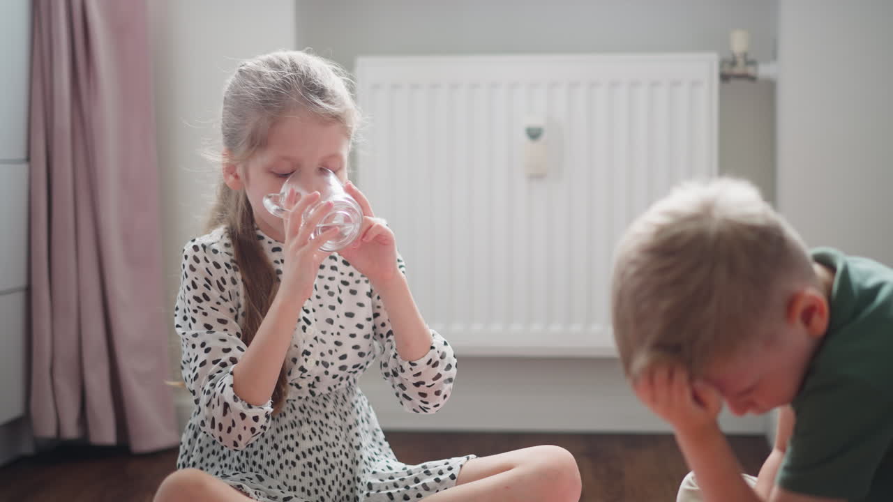 Little girl with long blond hair in spotted dress drinking water from clear glass cup while little boy in green shirt sits beside thoughtfully bent over