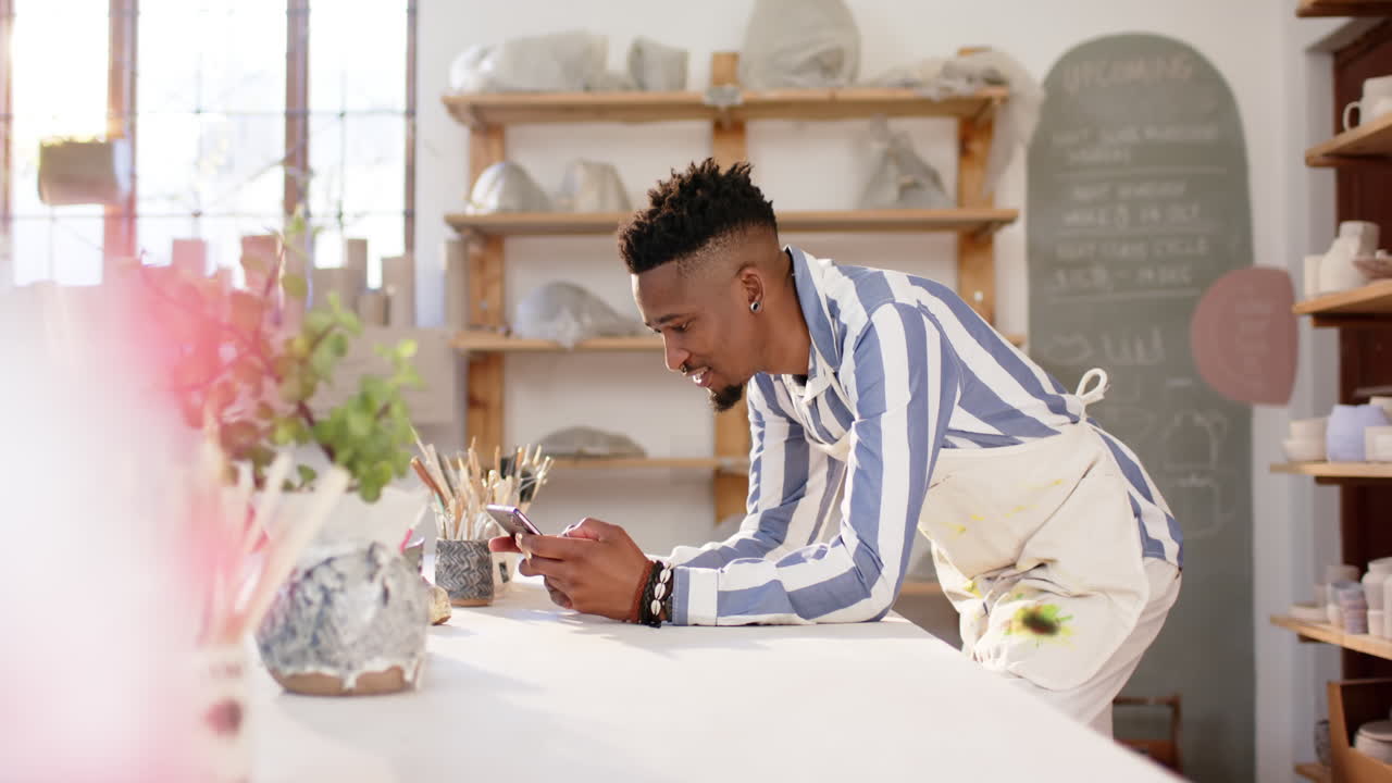 Happy african american man with beard using smartphone and smiling in pottery studio, slow motion
