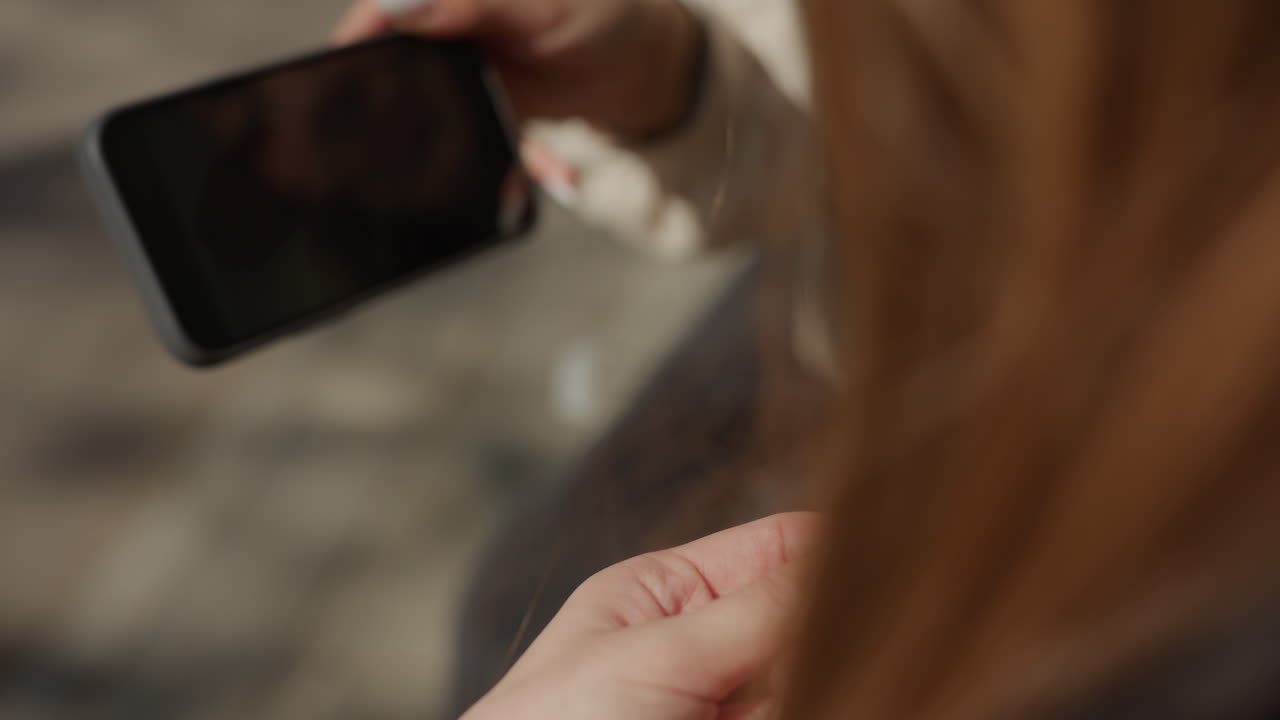 primer plano de una mujer asiática cortando hilo cerca de un teléfono; sus manos usan unas tijeras pequeñas para cortar los extremos del hilo; luz suave sobre la manicura; banco informal; ambiente relajado y tranquilo; detalle acogedor hecho a mano