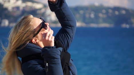 Una-Joven-Rubia-Pasa-Por-Su-Cabello-Y-Se-Quita-Las-Gafas-De-Sol,-El-Mar-Azul-En-Un-Enfoque-Suave-Detrás-De-Ella