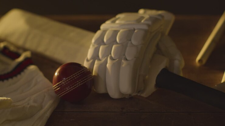 Natura morta di cricket con primo piano di mazza, palla, guanti, paletti, maglione e traversini che giacciono su una superficie di legno nello spogliatoio 2