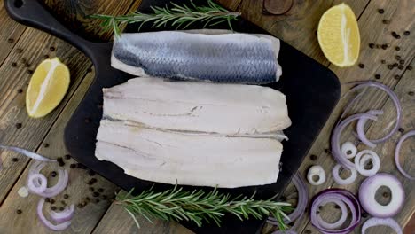 filete de arenque salado con cebollas, limón y romero en la tabla de corte oscura girando.