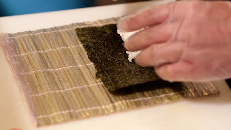 Chef-Puts-Seaweed-Wrap-And-Sushi-Rice-On-A-Bamboo-Mat-For-Rolling---close-up,-slow-motion