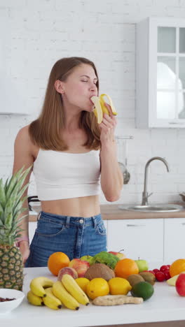 attractive model eating banana. healthy fruit and vegetables on the table. cheerful woman eats fruit in the modern light kitchen. vertical video