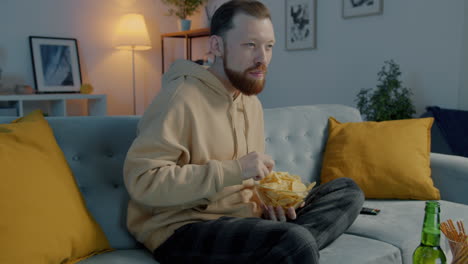 man watching tv at home, eating chips and drinking beer