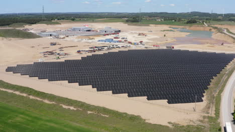solar panel array on sandy terrain near industrial site. wind turbines in distance. aerial pullback shot