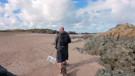 bald male carrying sand scoop walking dog on sunny rocky welsh beach active lifestyle exploring