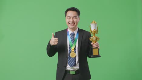 asian business man in a suit and tie with a gold medal and trophy showing thumbs up gesture and smiling to camera as the first winner on green screen background in the studio