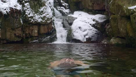 man plunges in cold water during ice swimming in subzero cold river by the beautiful waterfall in snowy mountains during winter day - static shot, slow motion