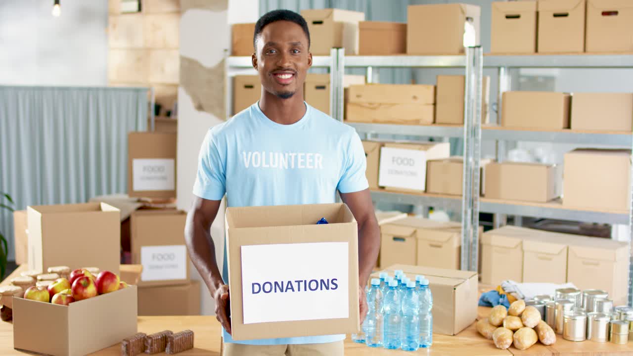 African american male volunteer holding donation box and smiling to the camera in charity warehouse