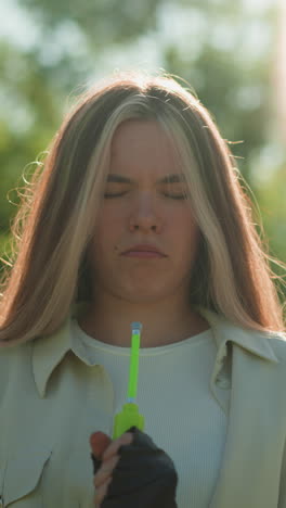 chica de pelo rubio de pie con una expresión pensativa, examinando cuidadosamente la boquilla de la bomba de aire verde contra el fondo de árboles exuberantes y iluminados por el sol