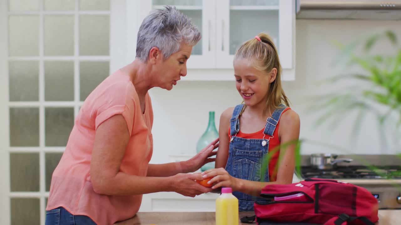 abuela caucásica en la cocina preparando un almuerzo empacado hablando con su nieta dándole fruta