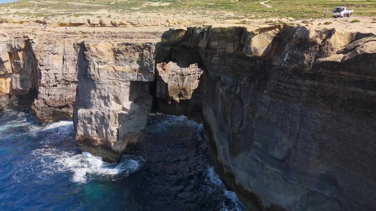 Wied il-Mielaħ in Gozo, Malta, natural sea arch and rugged coastal cliffs above blue Mediterranean water and waves along the shoreline, captured in aerial view.